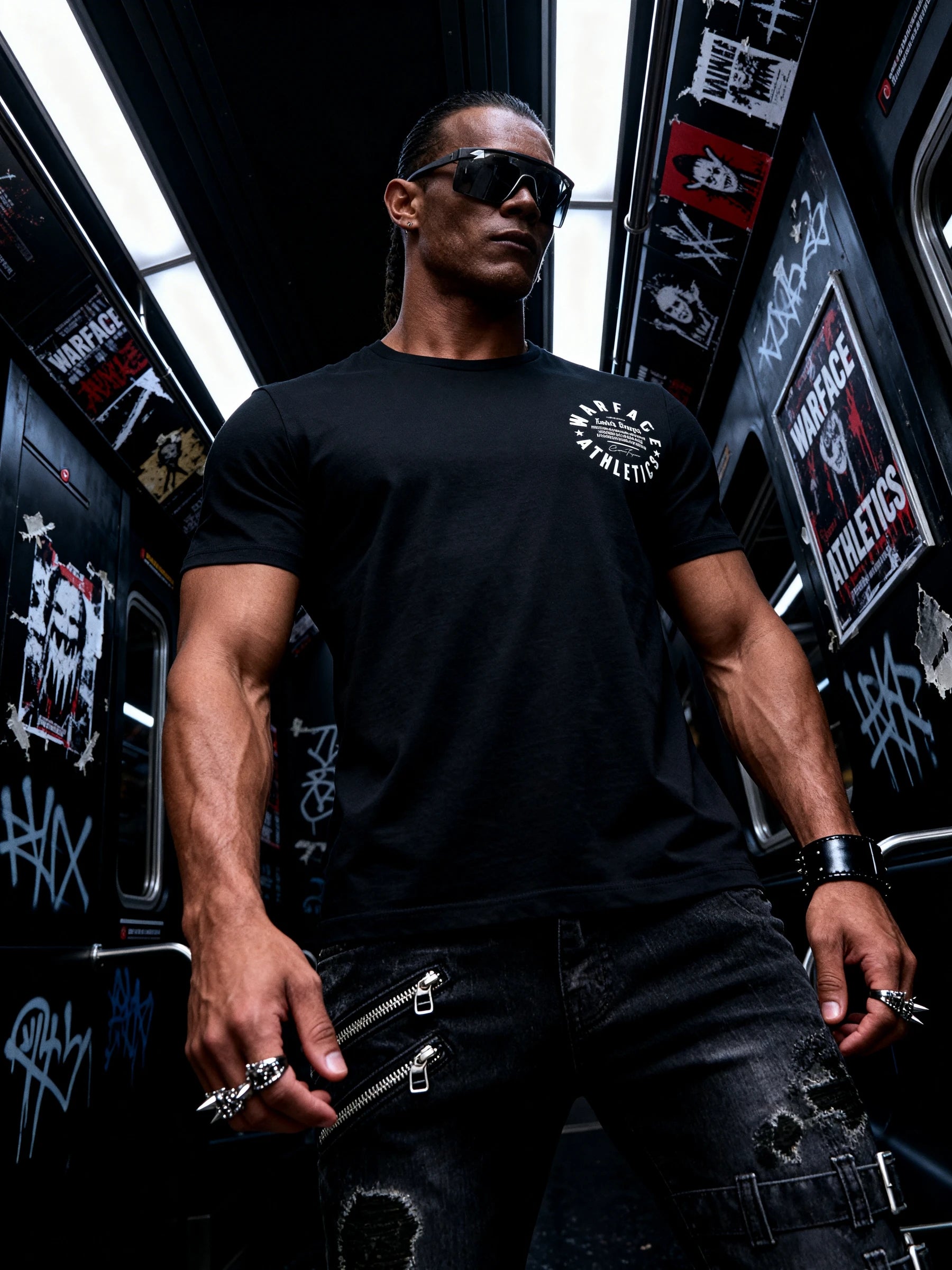 Man wearing a black gym t-shirt with graphics of Warface Athletics logo, sunglasses, and punk rings in a graffiti-covered subway train.